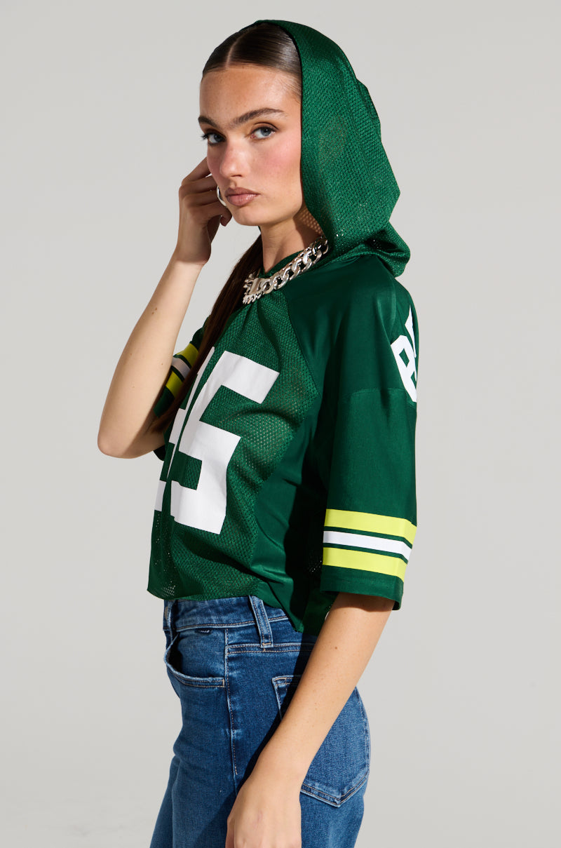 A woman dressed in a LUCKY JERSEY—a chic, sporty top crafted from semi-stretchy jersey fabric—displaying the number 15 and featuring a hood, combined with blue jeans. Her long, straight hair frames her face as she gazes directly at the camera, one hand gently touching her hair. A thick chain necklace adorns her look.