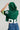 A person is pictured from behind wearing the green LUCKY JERSEY, a sporty chic top crafted from semi-stretchy jersey material and featuring the number 85 on the sleeve. They are adjusting its hood while paired with blue jeans, all set against a plain backdrop.