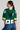 A person with long hair is wearing a green and white LUCKY JERSEY, numbered 95. The jersey features short sleeves and has a hood made of semi-stretchy jersey material. They are also wearing hoop earrings and blue jeans, standing against a plain background.