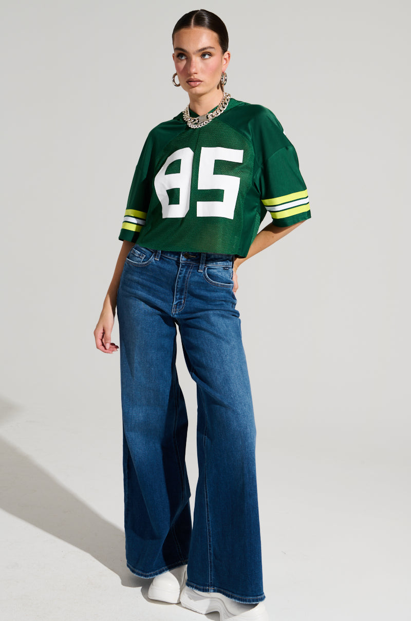 A person stands confidently, sporting their LUCKY JERSEY in semi-stretchy green material, numbered 95. It's paired with wide-leg blue jeans and white shoes. Accessories like a chunky metallic necklace and hoop earrings complete this sporty chic ensemble against a plain background.