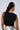 A person with long, dark hair styled in a high ponytail is facing away and wearing the "Do I Look Like I Care Tank," a sleeveless black crop top adorned with small rhinestone embellishments. They are also wearing large hoop earrings and metallic silver high-waisted pants.