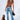 A woman stands with her back to the camera, showcasing her tattoos and a BUTTERFLY EFFECT SLEEVELESS KNIT BODYSUIT featuring crisscross straps. She pairs it with blue jeans that have embellished pockets and carries a black handbag. A smaller inset image from the front reveals cutouts on the sides of her bodysuit, highlighted by a Silver Butterfly Pendant.