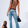 A woman stands with her back to the camera, showcasing her tattoos and a BUTTERFLY EFFECT SLEEVELESS KNIT BODYSUIT featuring crisscross straps. She pairs it with blue jeans that have embellished pockets and carries a black handbag. A smaller inset image from the front reveals cutouts on the sides of her bodysuit, highlighted by a Silver Butterfly Pendant.