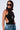 A person with dark hair wearing large, dark sunglasses, the BUTTERFLY EFFECT SLEEVELESS KNIT BODYSUIT in black with side cutouts, and blue denim jeans. They have a tattoo on their left shoulder and are accessorized with a long necklace featuring a silver butterfly pendant.