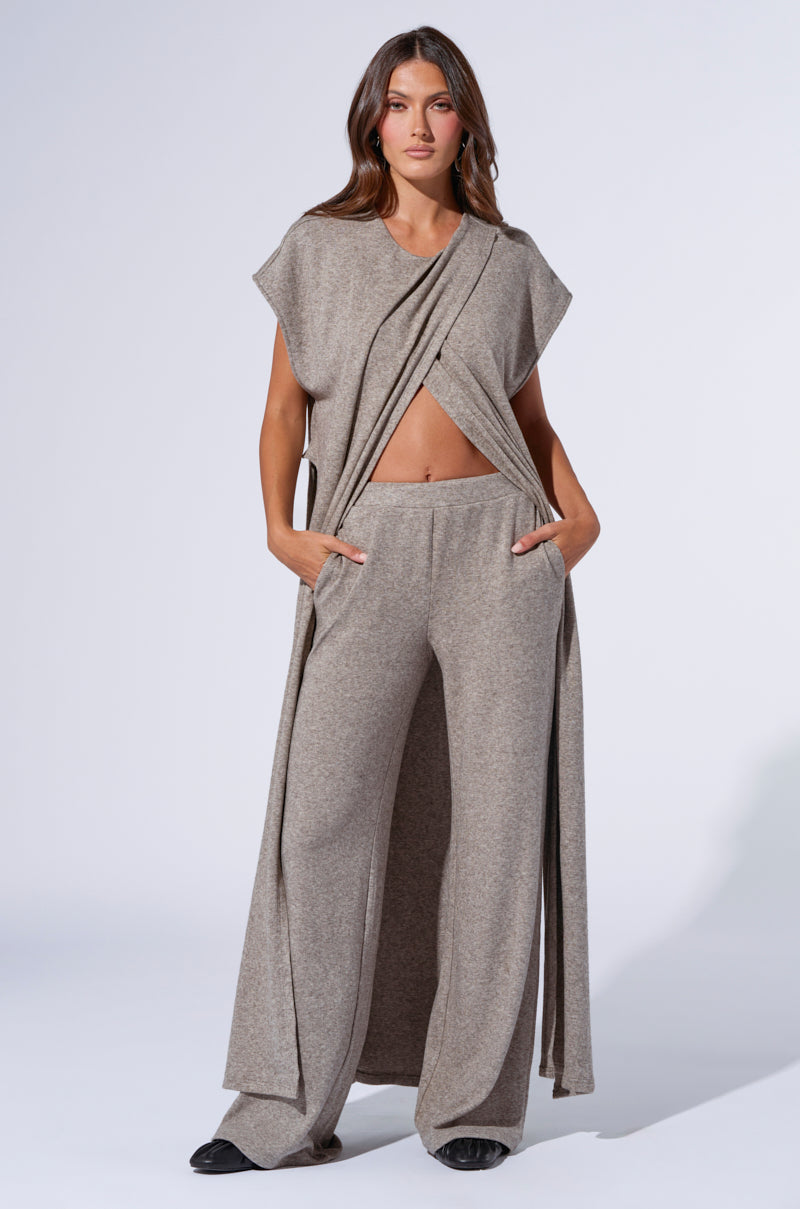 A woman stands against a plain background wearing a matching two-piece beige outfit. The top is sleeveless with draping fabric and asymmetric cuts, revealing a glimpse of her midriff, paired with high-rise relaxed fit wide-leg pants. She has her hands in her pockets while looking into the camera, dressed in what might be similar to the VIBE OUT FLARE LEG KNIT PANT IN GREY. 0