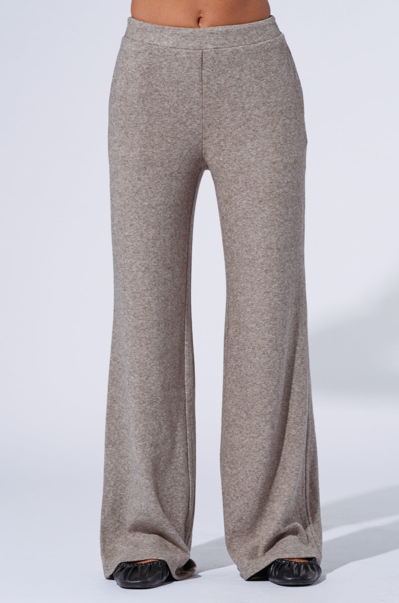 A person is standing wearing the VIBE OUT FLARE LEG KNIT PANT IN GREY, which are high-rise relaxed fit, wide-leg pants that extend to the ground. The knit material of the pants appears soft and comfortable. Only the lower half of their body, from the waist down, is shown. They are also wearing black shoes. 3