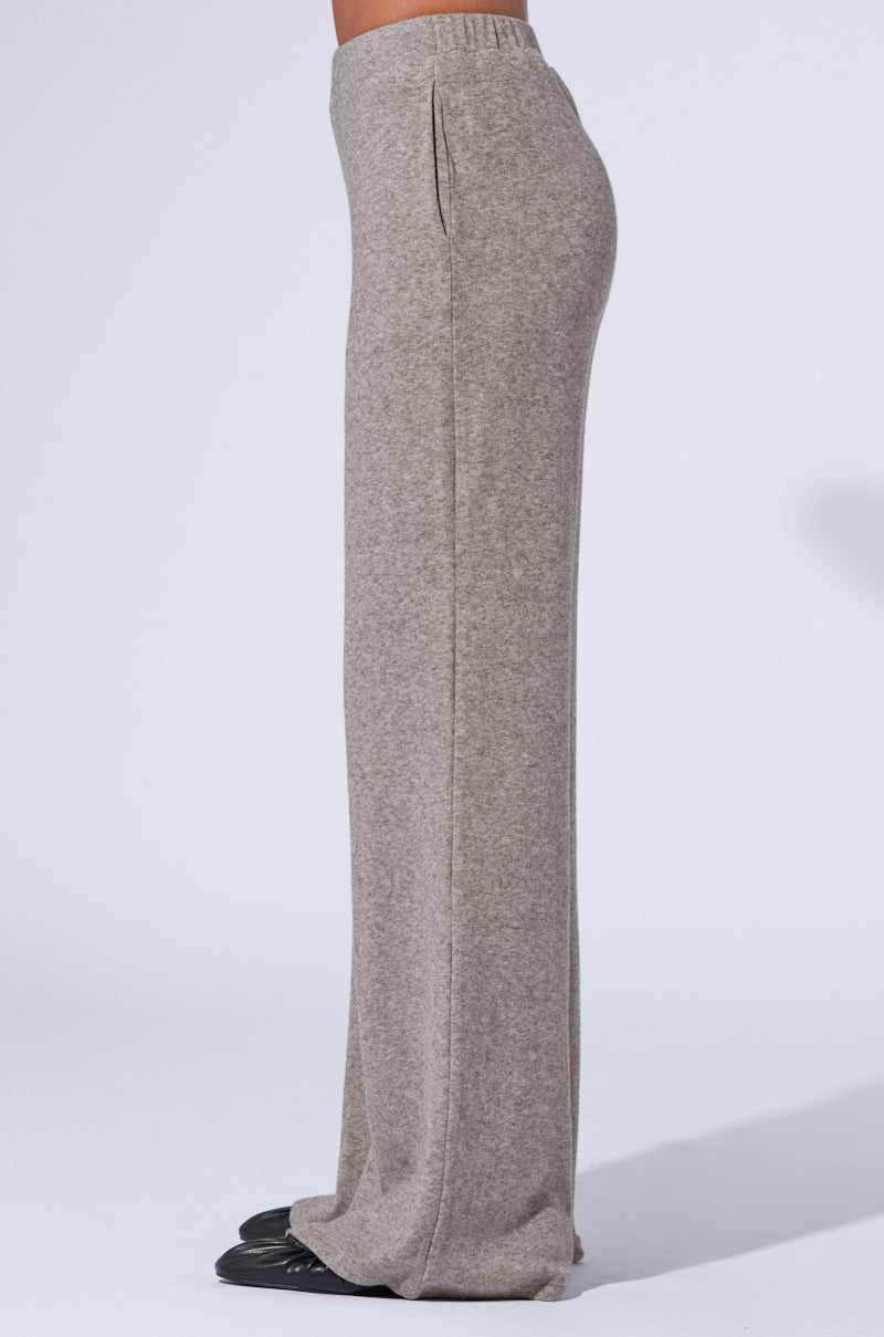 A person is wearing the VIBE OUT FLARE LEG KNIT PANT IN GREY. These high-waisted, wide-leg knit pants feature an elastic waistband and a soft, comfortable texture. With a loose fit that extends to cover the shoes, only the lower half of the body is visible against a plain background. 4