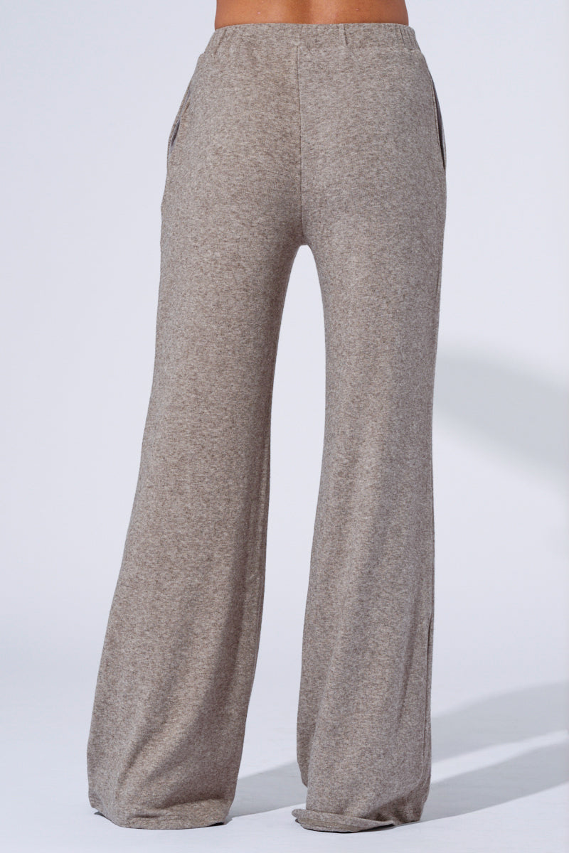 A person is shown from the back, wearing grey, high-rise relaxed fit knit pants called the Vibe Out Flare Leg Knit Pant in Grey. The pants have a flare leg and pockets on both sides. The plain white background draws focus to the comfortable loungewear style of the pants. 5