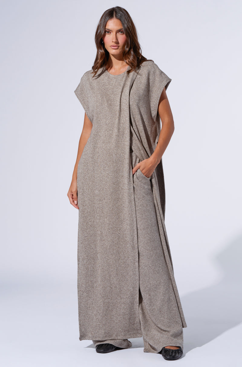 A woman stands against a plain background wearing the VIBE OUT FLARE LEG KNIT PANT IN GREY. The pants are long and loose-fitting with an elegant drape. She pairs the grey knit pants perfectly with black shoes, and her hair is styled in loose waves, emphasizing the effortless comfort of versatile loungewear. 6
