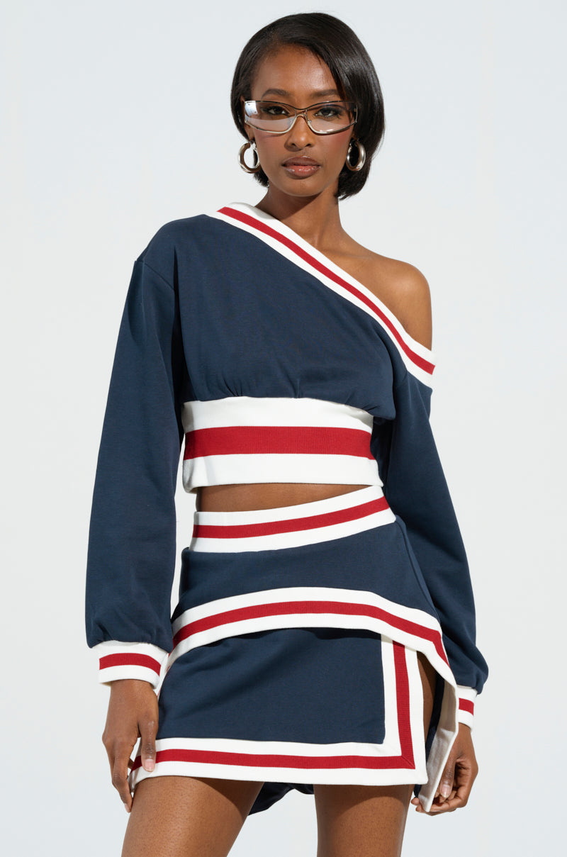 A person wearing a stylish MAEVE Off-The-Shoulder Cropped Sweatshirt in navy, featuring white and red stripes, stands confidently. The ensemble includes a layered skirt and is accessorized with glasses and hoop earrings.