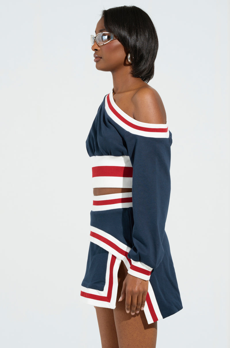 A woman with short dark hair is wearing glasses and a striking navy off-the-shoulder cropped sweatshirt, echoing the style of the MAEVE OFF THE SHOULDER CROPPED SWEATSHIRT IN NAVY. She stands gracefully in profile against a light background.