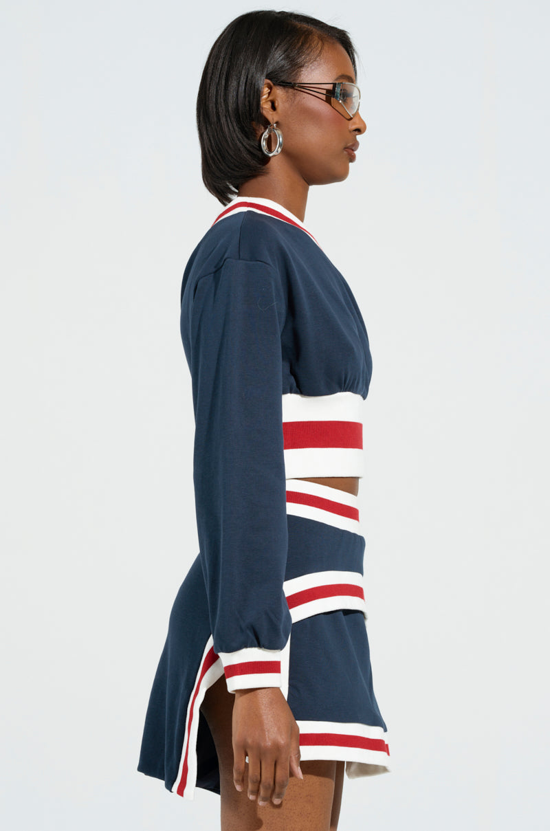 A woman stands in profile against a light background, wearing the AKIRA Label MAEVE Off The Shoulder Cropped Sweatshirt in Navy, along with a matching skirt set featuring white and red stripes. Her short dark hair is complemented by hoop earrings and glasses, while the off-the-shoulder design adds a touch of flair.