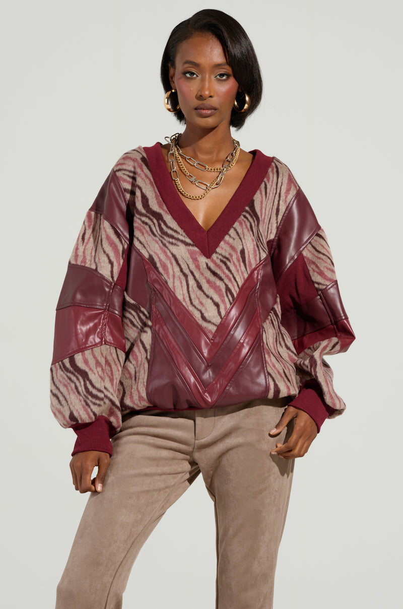 A woman models the stylish WILD SIDE MIXED MEDIA SWEATSHIRT, showcasing its zebra pattern and leather accents. She pairs it with tan suede pants and accessorizes with gold hoop earrings and layered necklaces, standing against a plain background.