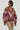 A person wearing a WILD SIDE MIXED MEDIA SWEATSHIRT featuring maroon leather accents and zebra-like patterns, paired with light brown pants. They have short black hair and are accessorized with layered gold necklaces. The background is plain white.
