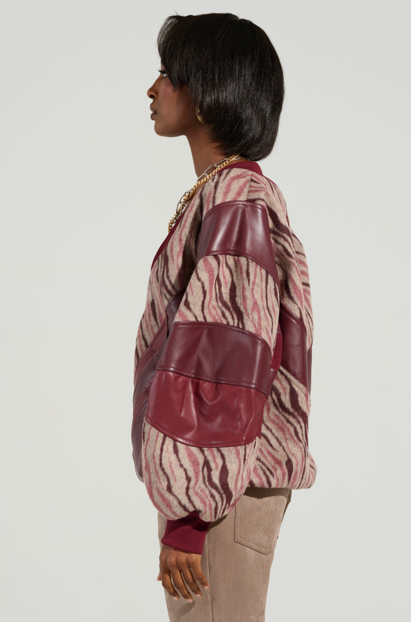 A person in profile is wearing the WILD SIDE MIXED MEDIA SWEATSHIRT, featuring a stylish pink and brown zebra print combined with maroon leather patches. This zebra-striped fleece offers a unique, oversized design. The individual has short, dark hair and pairs the sweatshirt with light brown pants against a plain background.