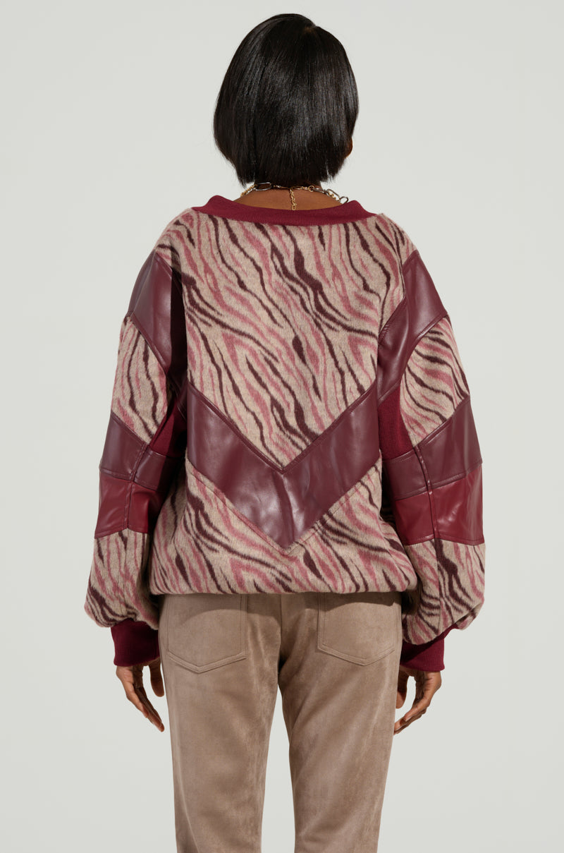 A person is seen from behind wearing the WILD SIDE MIXED MEDIA SWEATSHIRT, which combines zebra-striped fleece with dark red leather patches, along with beige pants. The daring textures and colors evoke a wild side vibe as they stand against a plain background.