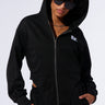 Person wearing the TAKE THE LONG WAY OVERSIZED ZIP UP HOODIE from the AKIRA Label, a black oversized zip-up made with a slight stretch cotton fabric and the hood up, paired with black sunglasses. Holding the pockets of their hoodie, which features a small white design on the left chest area. Curly hair against a plain white background.