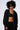 A person with curly hair is wearing dark sunglasses, a black AKIRA Label crop top, and black pants. They are standing against a light background and looking forward. The midriff is revealed as their TAKE THE LONG WAY OVERSIZED ZIP UP HOODIE hangs open, unzipped.