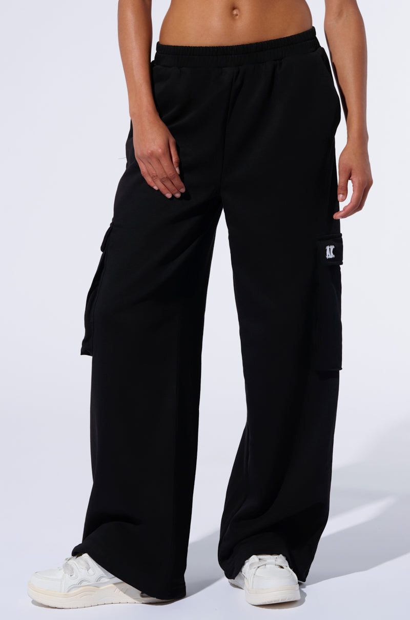 A person is wearing the black, loose-fitting TAKE THE LONG WAY CARGO JOGGER pants with cargo flap pockets and white sneakers. The image is cropped just above the waist, showcasing the relaxed fit of these joggers from AKIRA Label. One hand is resting in a pocket against a plain, light-colored background.