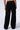 A person is facing away, wearing the TAKE THE LONG WAY CARGO JOGGER from the AKIRA Label. These black cargo pants feature two large side flap pockets on each leg, fit loosely, and extend down to their feet. The plain, light-colored background highlights only their lower back and legs.