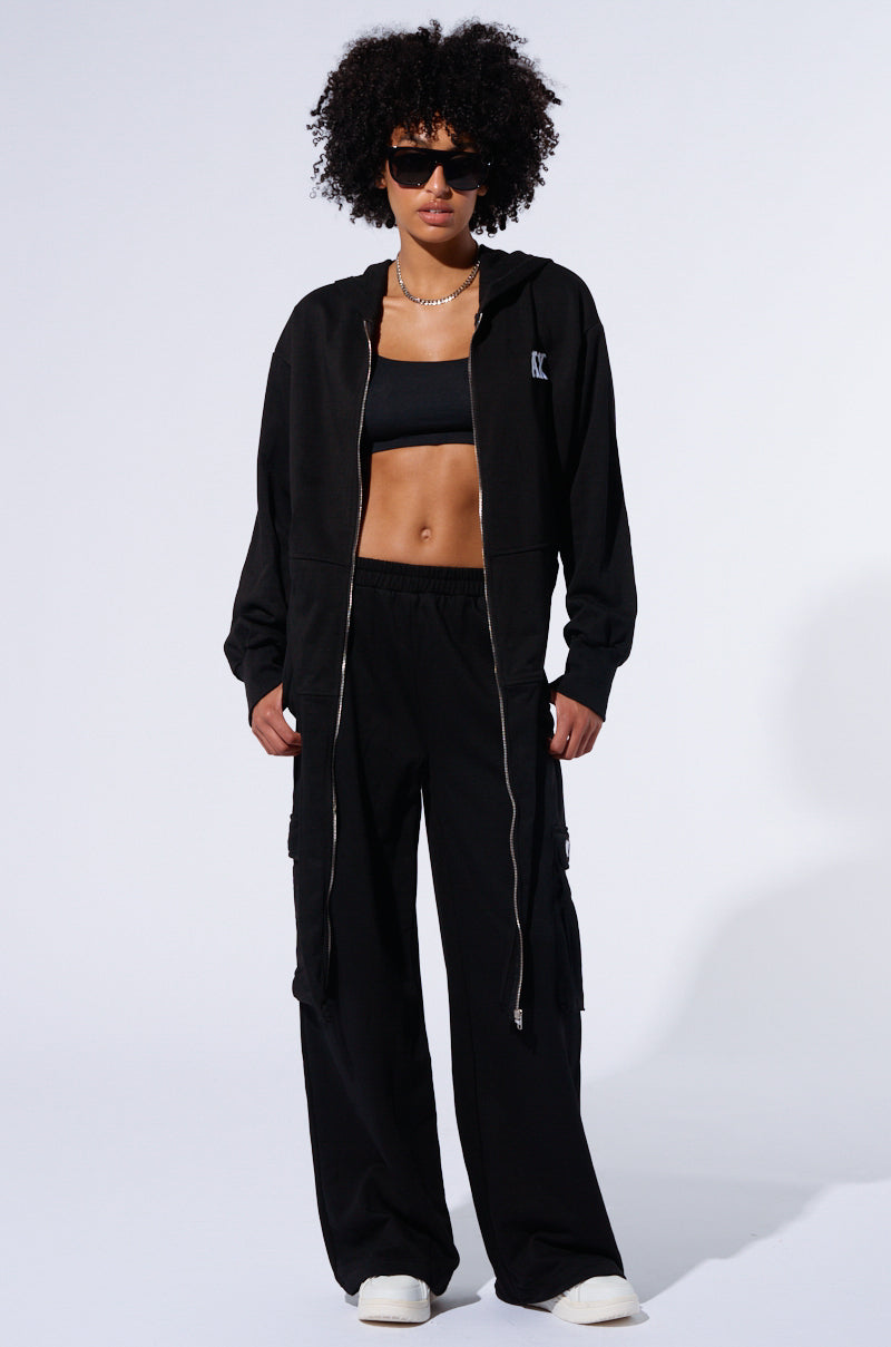 A person with curly hair is standing against a white background, wearing a black zip-up hoodie, a black sports bra, Take The Long Way Cargo Jogger pants with cargo flap pockets, and white shoes. They are also wearing sunglasses and a pearl necklace.