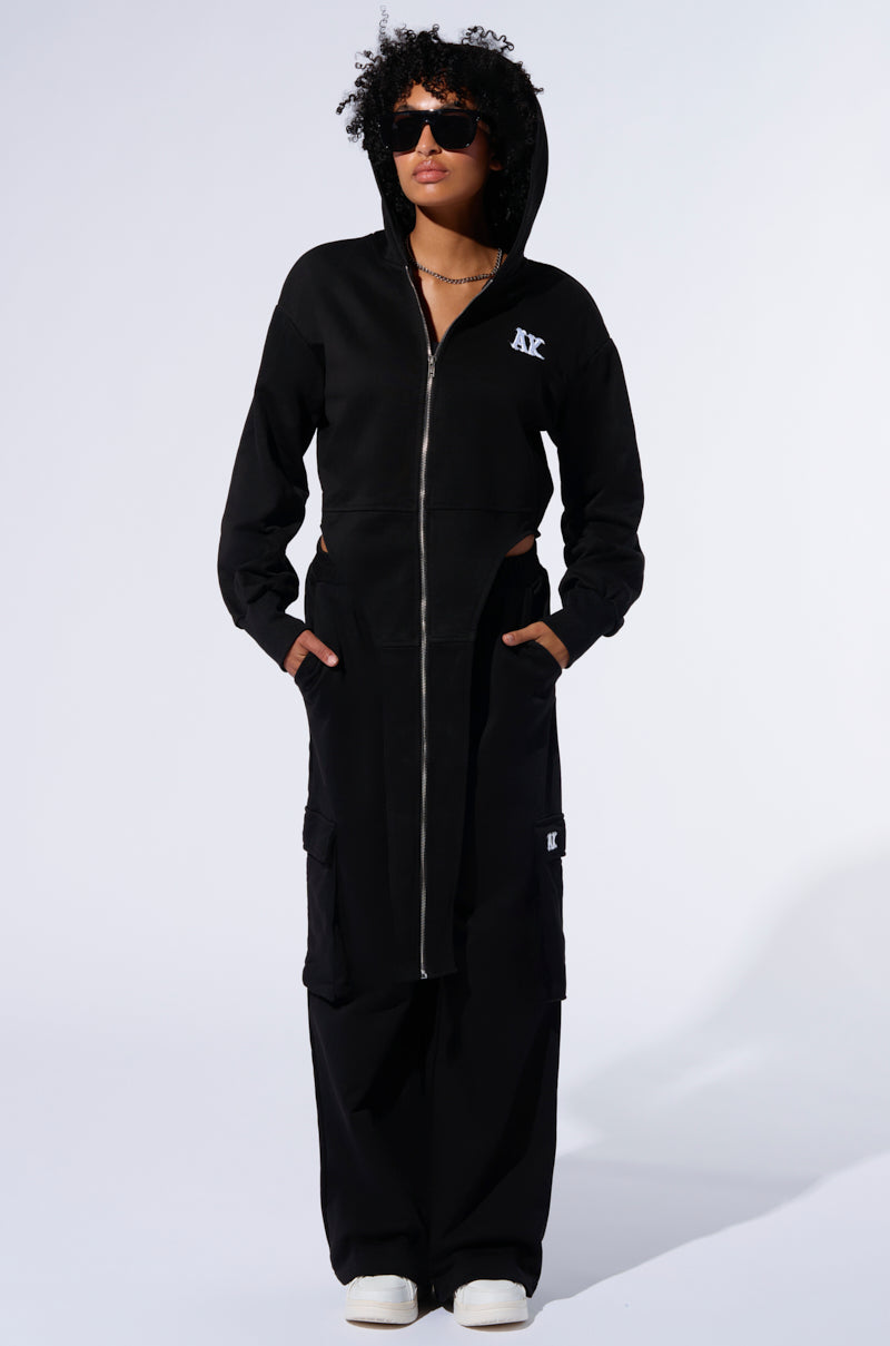 A person with curly hair and sunglasses poses in a black, full-length hooded coat with a zip-up front. Their hands rest in the coat's pockets, and they are dressed in AKIRA Label’s loose-fitting TAKE THE LONG WAY CARGO JOGGER pants featuring cargo flap pockets, paired with white sneakers against a plain white background.