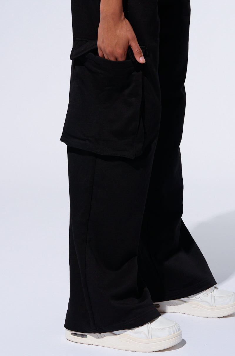 A person stands against a light background, wearing black TAKE THE LONG WAY CARGO JOGGER pants with cargo flap pockets on the side and white sneakers. The person’s hand is partially visible, casually resting on top of the pocket of their joggers. The lower half of their body is shown.
