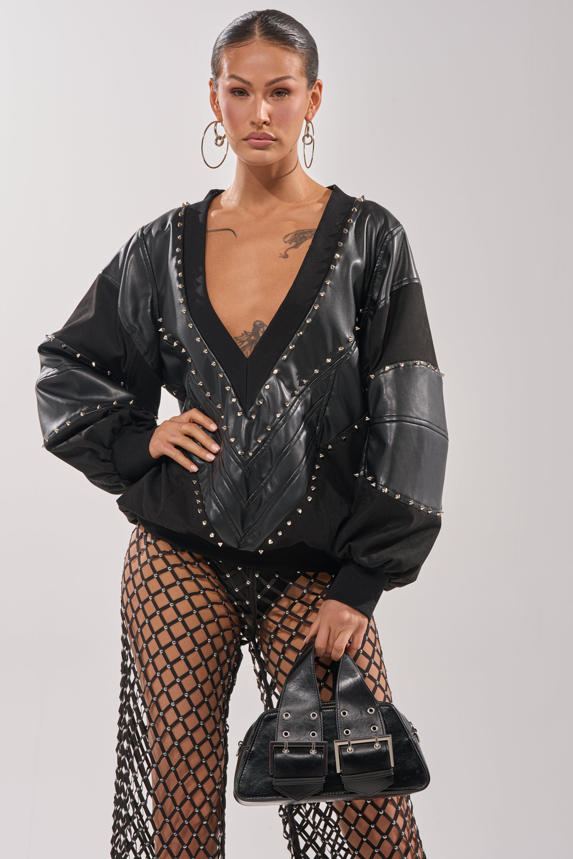 A model poses confidently in the LETS DO THIS FAUX LEATHER SWEATSHIRT, styled with black mesh fishnet pants, hoop earrings, and a black handbag with silver buckles, showcasing bold streetwear against a plain background. 0