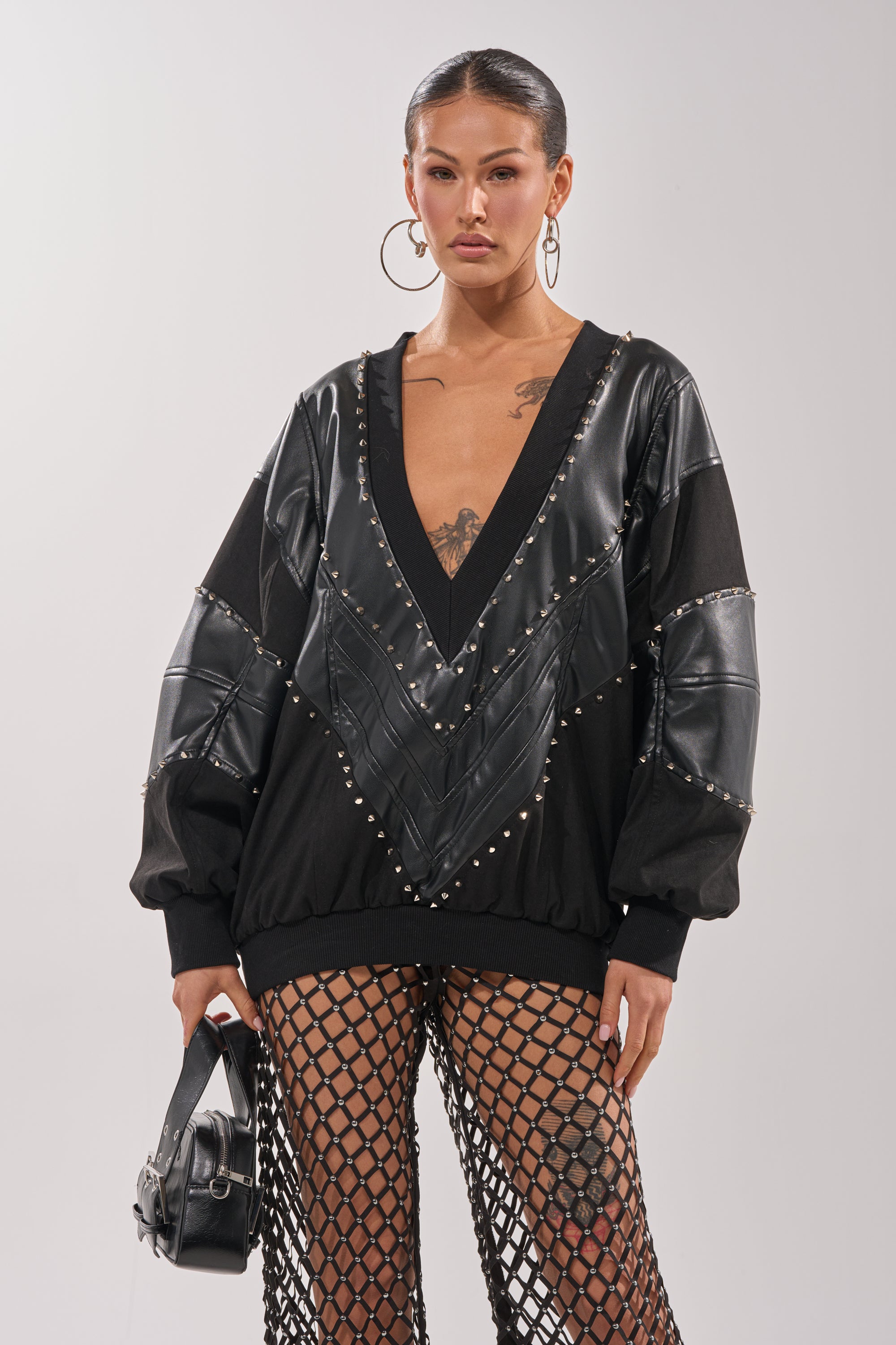 A woman wearing the LETS DO THIS FAUX LEATHER SWEATSHIRT with studs, large hoop earrings, mesh fishnet pants, and a small black bag stands against a plain light background. 1