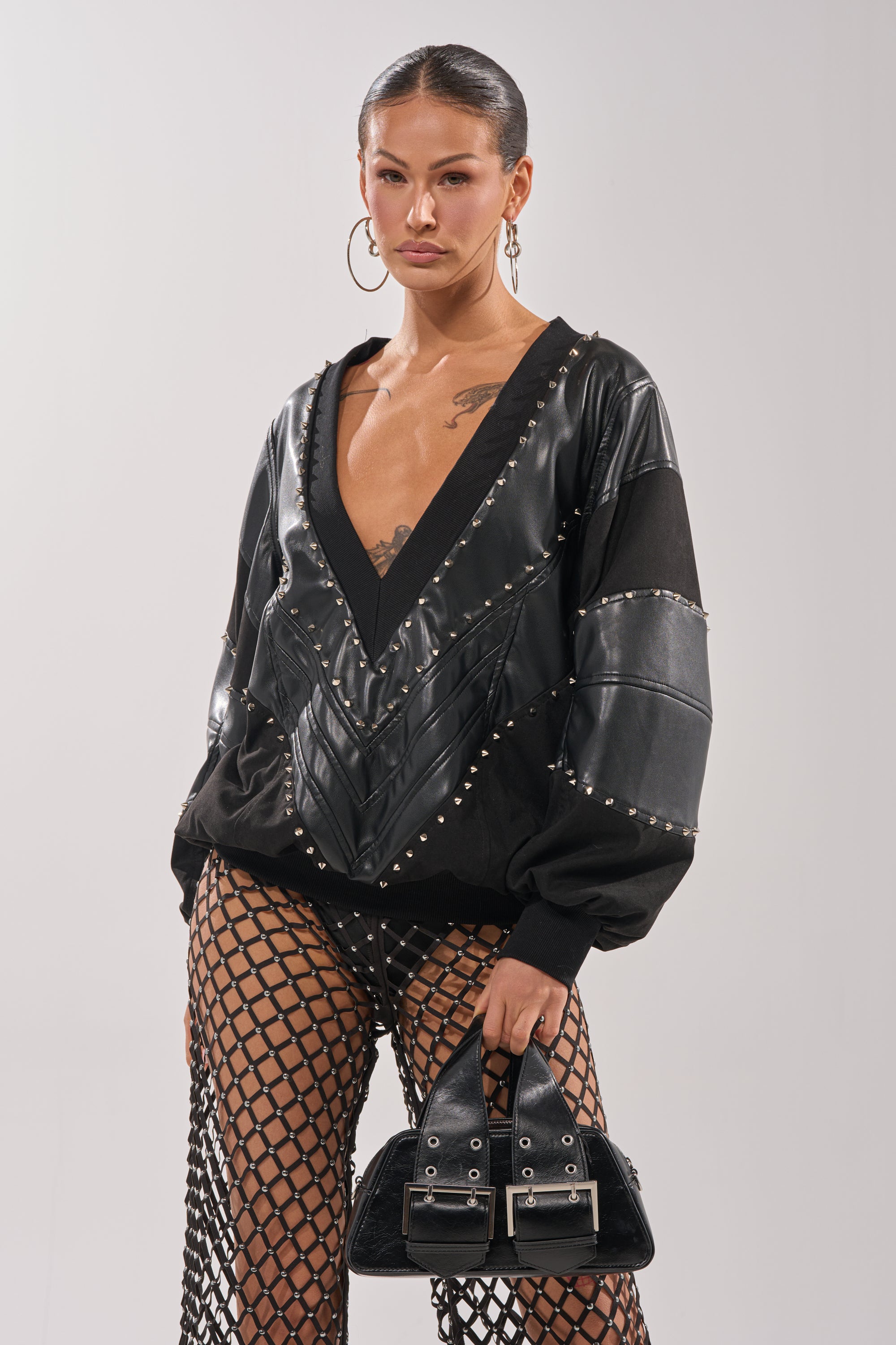 A woman in the LETS DO THIS FAUX LEATHER SWEATSHIRT with a deep V-neck, studded details, fishnet pants, and hoop earrings holds a black handbag, showcasing bold streetwear style against a light gray background. 2