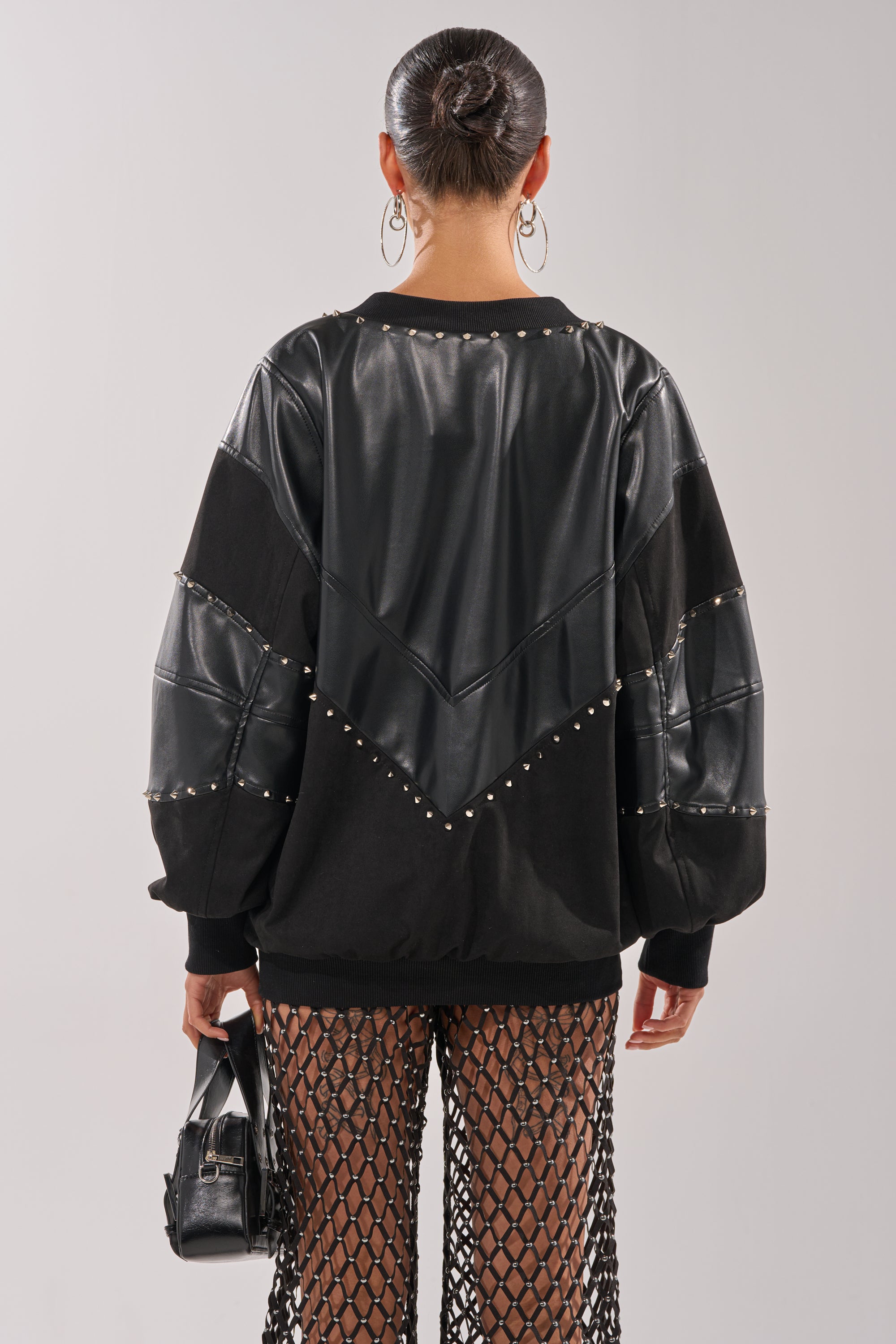 A person with a slick bun faces away, wearing the LETS DO THIS FAUX LEATHER SWEATSHIRT with metal studs, black mesh pants, hoop earrings, and holding a small black handbag. 5