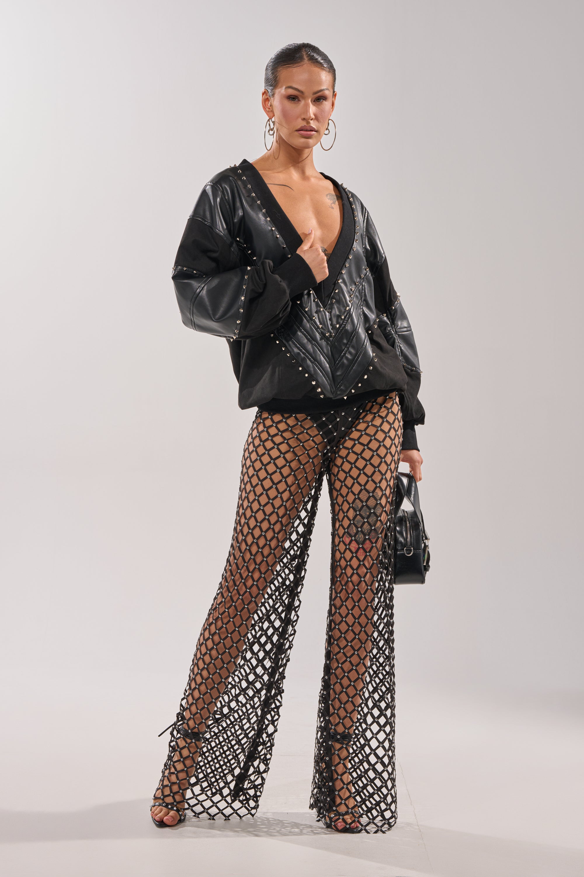 A woman stands confidently in the LETS DO THIS FAUX LEATHER SWEATSHIRT styled with wide fishnet pants over briefs and heels, accessorized with hoop earrings, a small handbag, and her hair slicked back for a bold streetwear-inspired look. 6