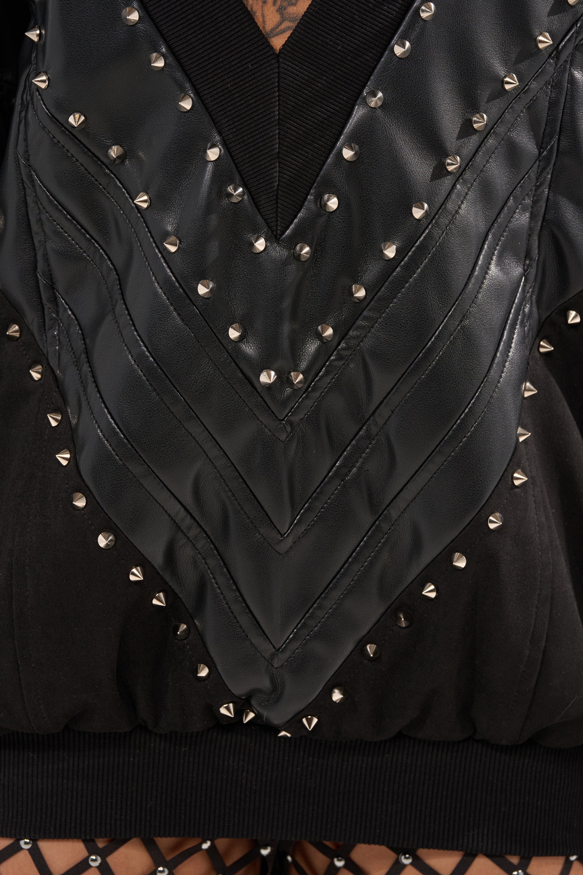 Close-up of the LETS DO THIS FAUX LEATHER SWEATSHIRT, a black streetwear jacket with shiny chevron-quilted panels, silver stud details, ribbed hem, and a glimpse of a fishnet layer underneath. 7