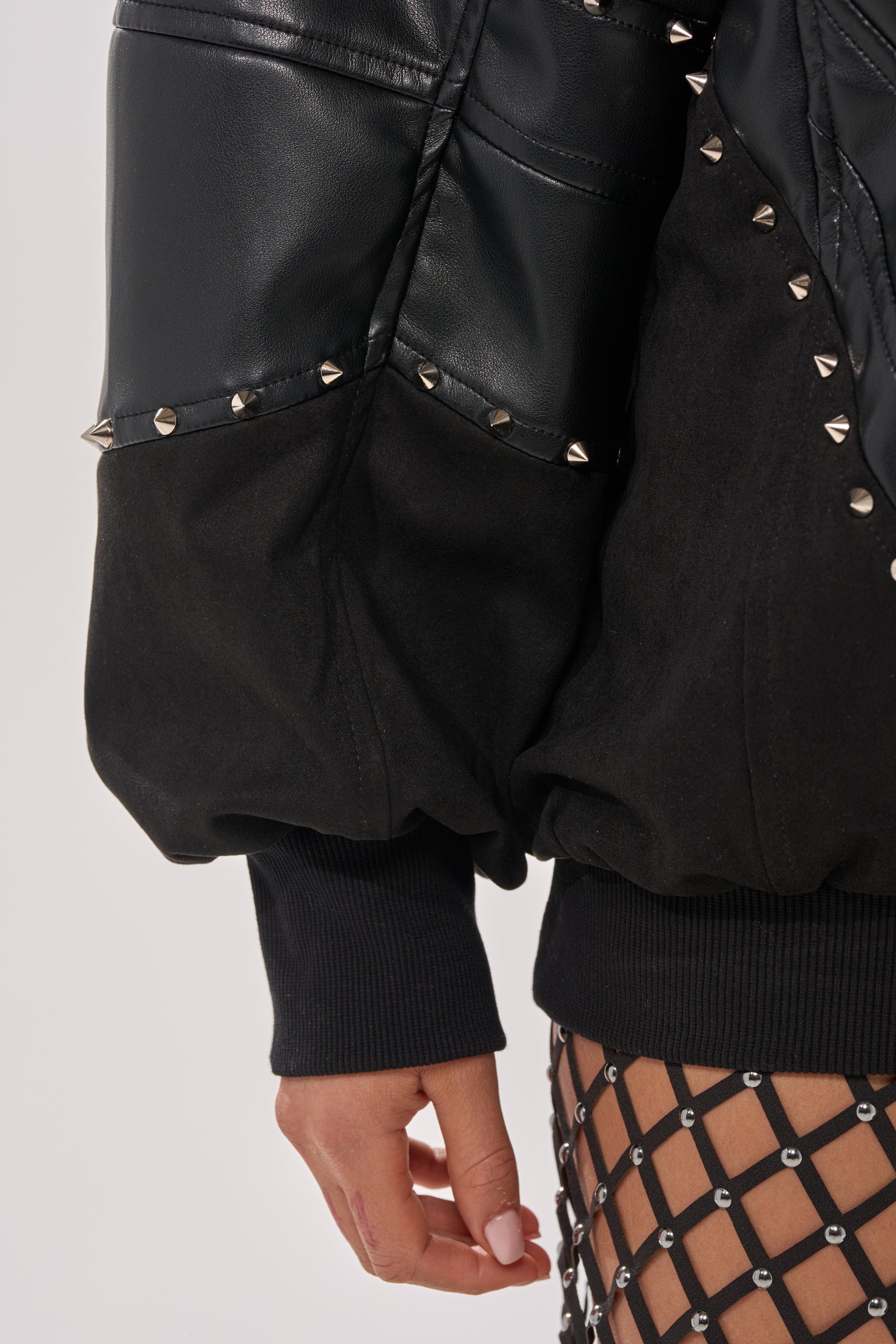 Wearing the LETS DO THIS FAUX LEATHER SWEATSHIRT with chevron-quilted panels, metallic studs, black ribbed cuffs, and a fishnet-patterned skirt—bold streetwear style. Only their hand and part of the outfit are visible. 8