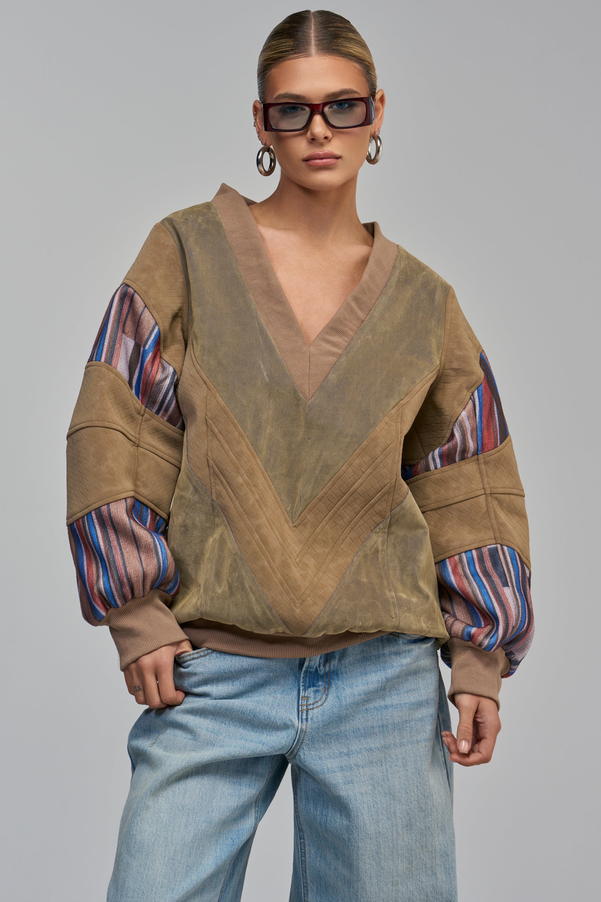 A woman wears THE NEW CLASSIC VELVET DENIM CHEVRON SWEATSHIRT, a brown V-neck streetwear top with chevron paneling and striped sleeve accents, paired with light-wash denim.