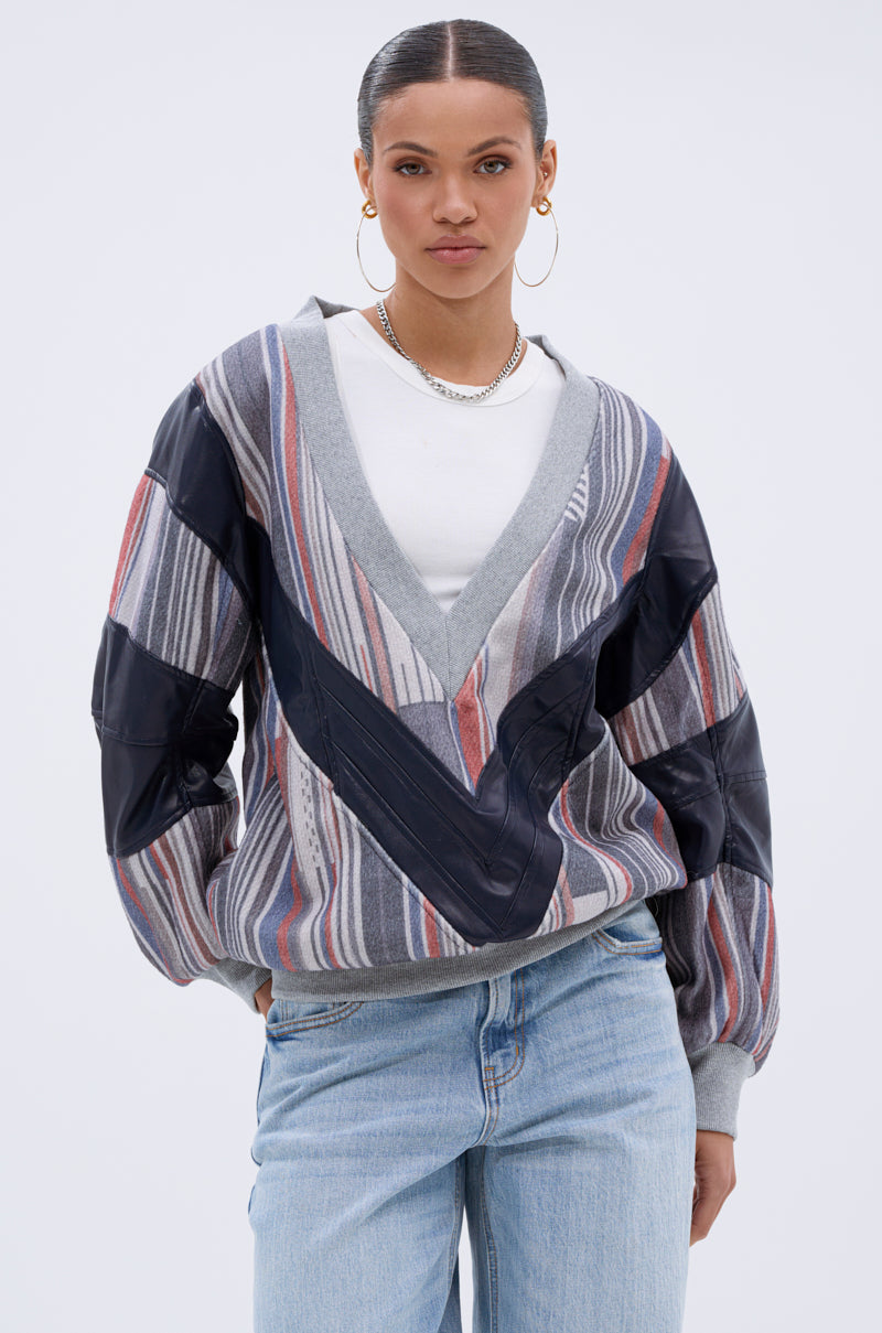 A woman with slicked-back hair, hoop earrings, a white t-shirt, and light blue jeans stands with one hand in her pocket, wearing the INTO THE WILD DEEP V SWEATSHIRT—an oversized gray, black, and red striped piece with leather accents.