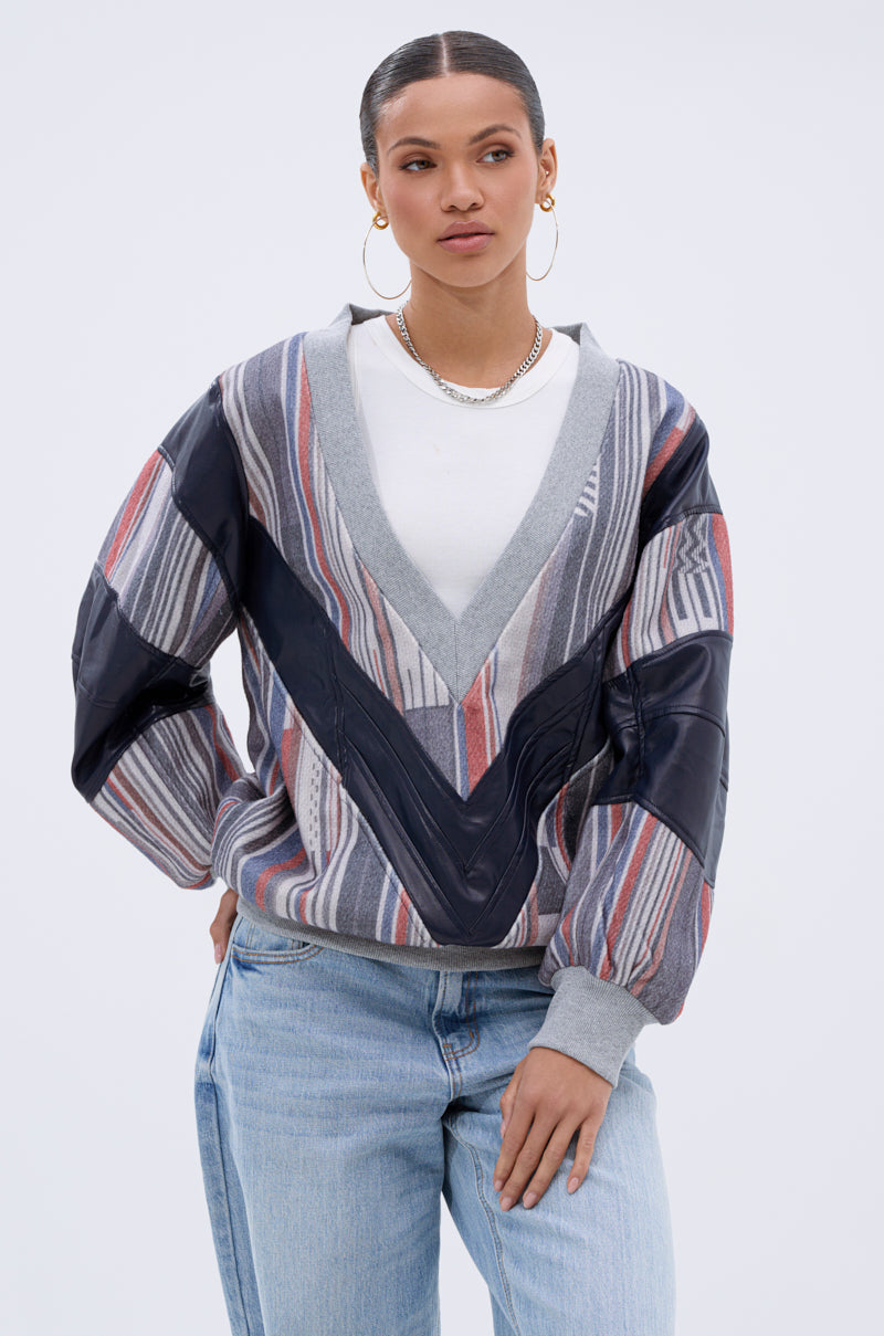 A woman stands with her hand in her pocket, wearing the INTO THE WILD DEEP V SWEATSHIRT over a white shirt. She accessorizes with hoop earrings and a chain necklace, hair pulled back, against a plain light gray background.