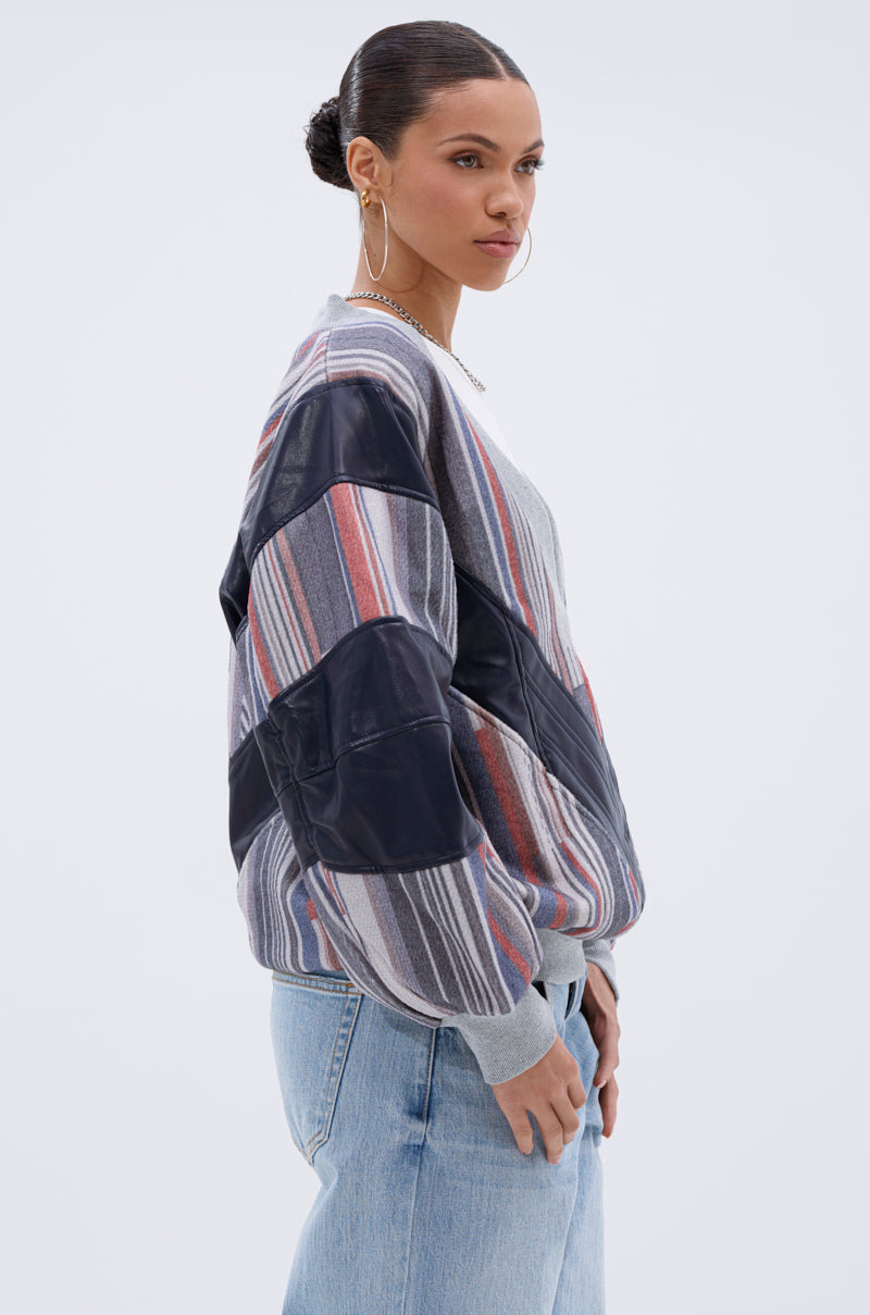 A woman with her hair in a bun, wearing large hoop earrings and light blue jeans, stands in profile against a plain background, modeling the INTO THE WILD DEEP V SWEATSHIRT featuring chevron stripes and a color-blocked design.