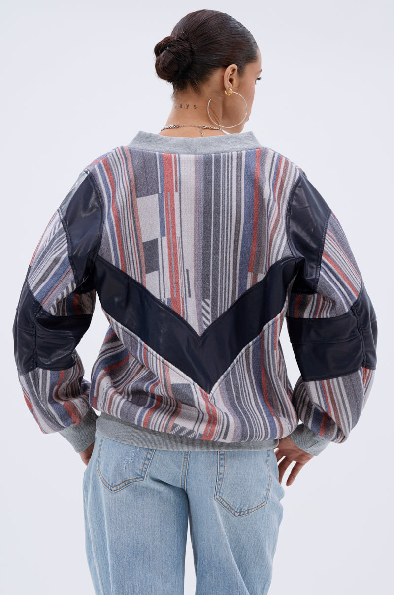 A woman with her hair in a bun and large hoop earrings wears the INTO THE WILD DEEP V SWEATSHIRT—a striped, Southwestern-inspired pullover with navy and gray accents—and light blue jeans, facing away to show the back of her outfit.