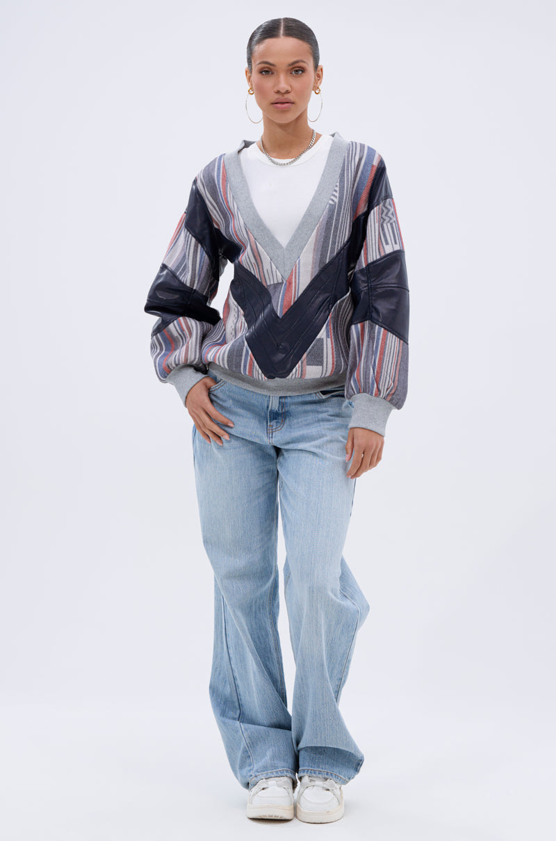 A woman stands facing forward against a plain light background, wearing hoop earrings, a necklace, light blue jeans, white sneakers, and the INTO THE WILD DEEP V SWEATSHIRT—a Southwestern pullover with blue, gray, and red tones.