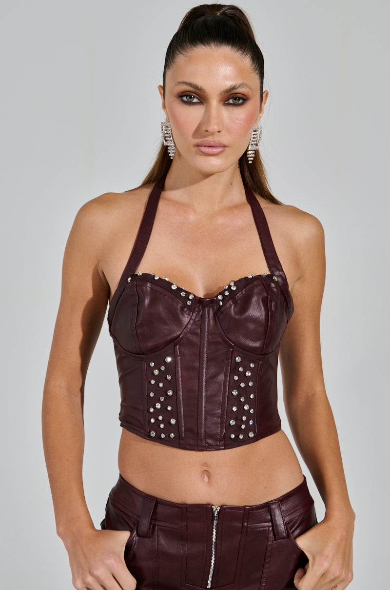 A confident person wears the TEMPTATION'S BEST FRIEND CORSET in burgundy, paired with matching pants. Their styled, long hair is pulled back, accessorized with large earrings against a neutral background.