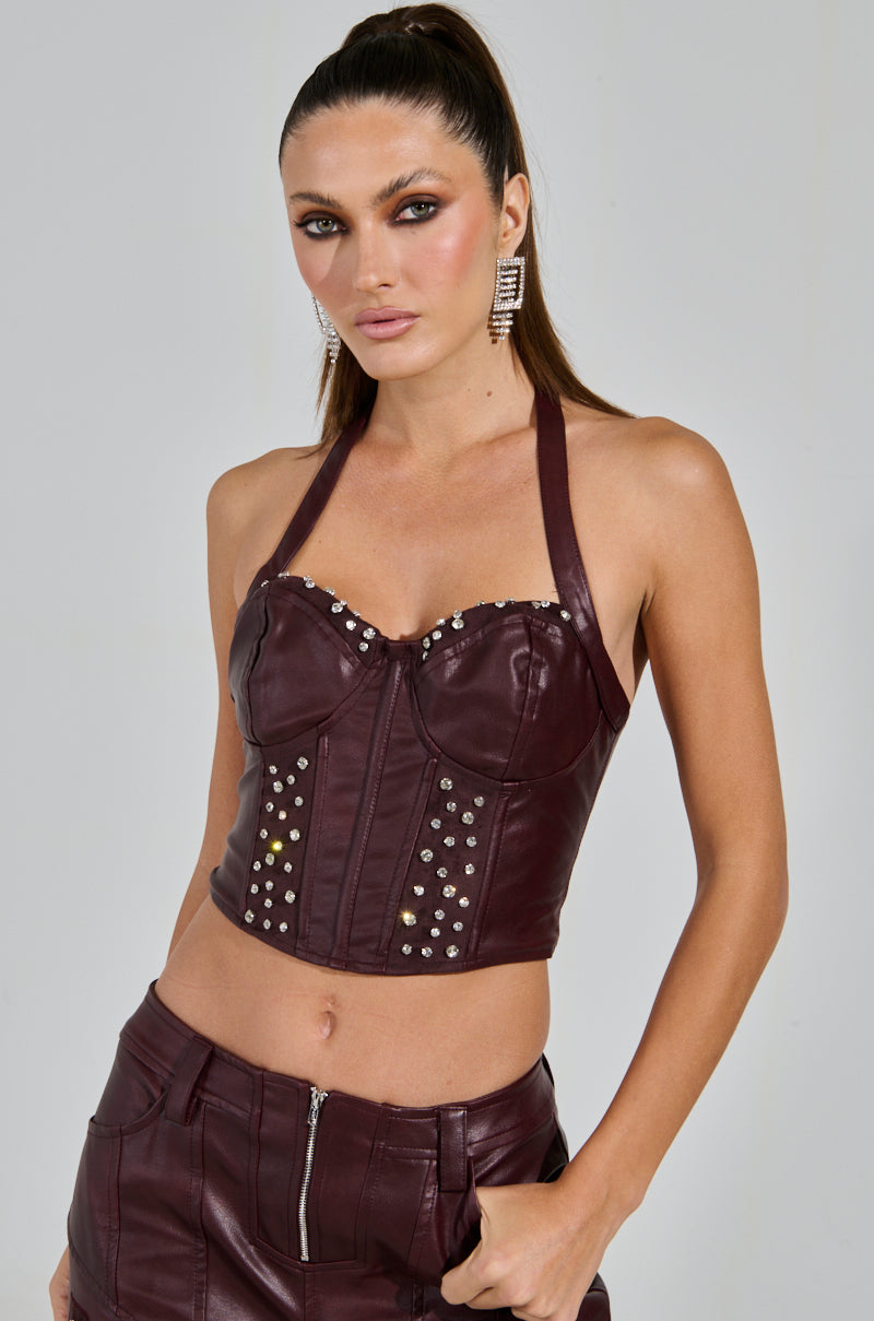 A woman poses confidently in the "Temptation's Best Friend Corset" in burgundy, embellished with studs, paired with matching leather pants. Her sleek, pulled-back hair and sparkling earrings enhance her chic, western-inspired look against a plain gray background.