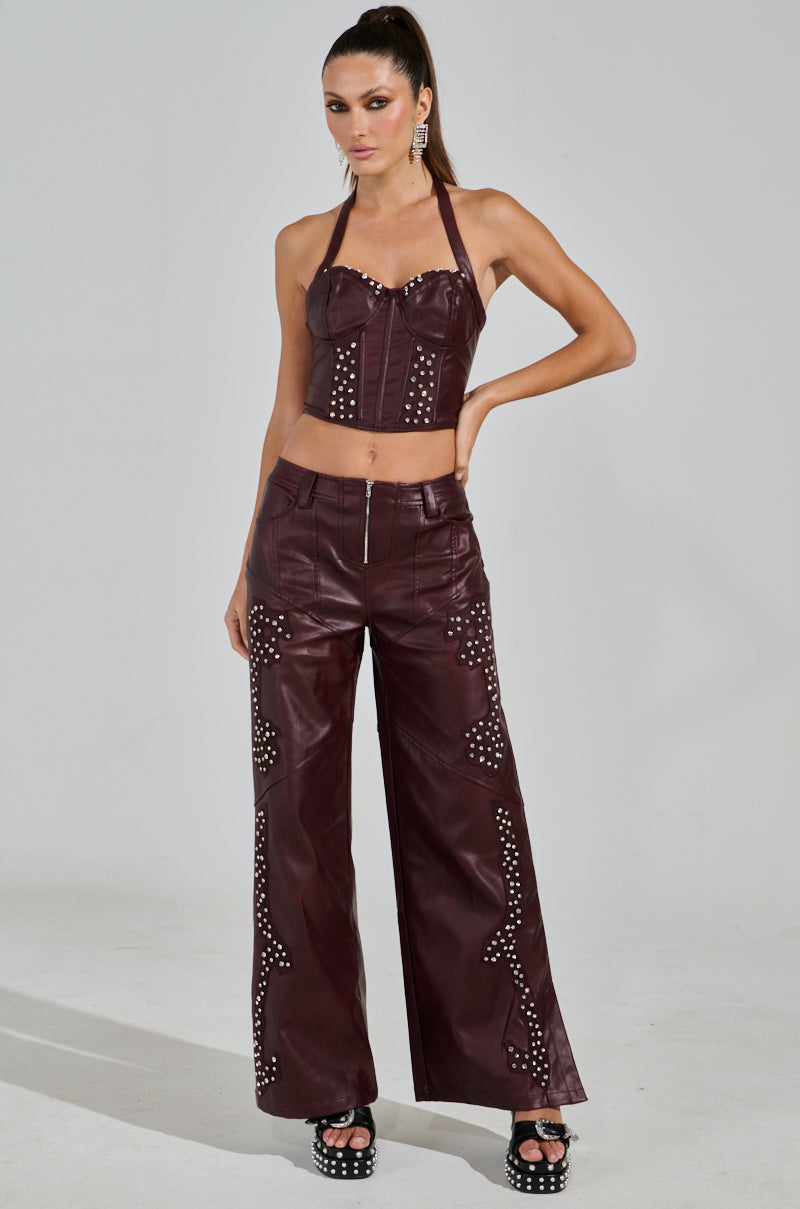 A person poses confidently in a western-inspired outfit, featuring the TEMPTATION'S BEST FRIEND CORSET IN BURGUNDY and matching wide-leg pants with silver studs. Their long hair is tied back, complemented by platform sandals and dangling earrings.
