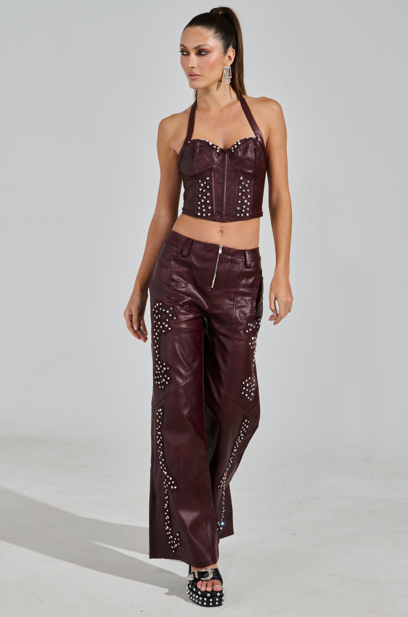 A person confidently wears the TEMPTATION'S BEST FRIEND CORSET in burgundy and pants with silver studs, paired with black platform heels and a sleek ponytail. They complete the western-inspired look against a plain gray background.