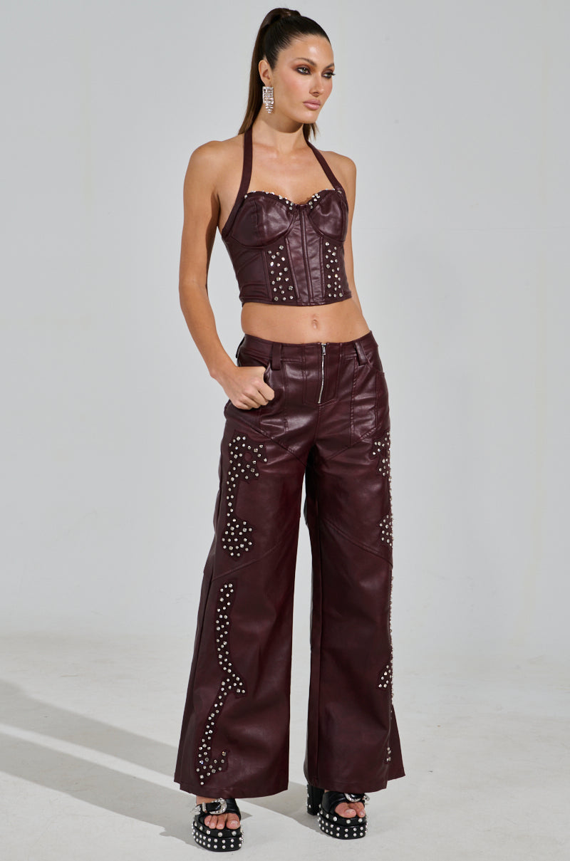 A woman poses in a western-inspired look, flaunting the Temptation's Best Friend Corset in burgundy leather, complemented by matching wide-leg pants with silver studs. Her long hair is tied in a high ponytail, and she wears black platform sandals. The simple gray backdrop highlights her striking ensemble.