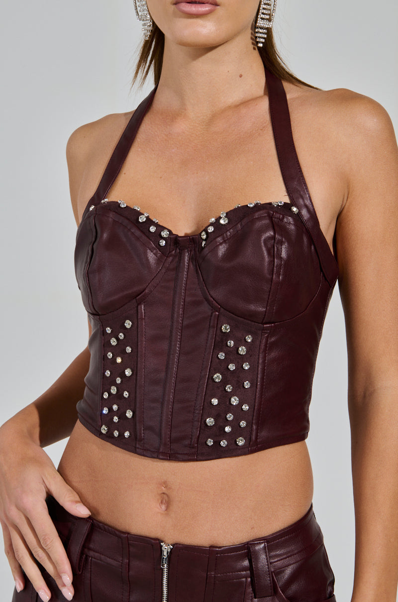 Wearing the TEMPTATION'S BEST FRIEND CORSET IN BURGUNDY—a dark maroon faux leather piece with rhinestone bust and front panel embellishments—a person strikes a western-inspired pose, paired with earrings, confidently resting one hand on their hip.