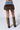 A Y2K baddie is seen from behind, sporting the FRANCESCA FUR TRIM DENIM MINI SKIRT that combines a blue denim waistband with a brown fur bottom. Their look is completed with tall brown snakeskin-patterned boots rising just below the knees. The background is plain light gray.