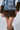 A Y2K baddie wearing the Francesca Fur Trim Denim Mini Skirt, a stretchy blue denim outfit adorned with brown faux fur accents on the skirt and sleeves. The mini skirt features belt with grommets and buckle detailing. The person is standing against a plain background, showcasing only their lower torso and legs.