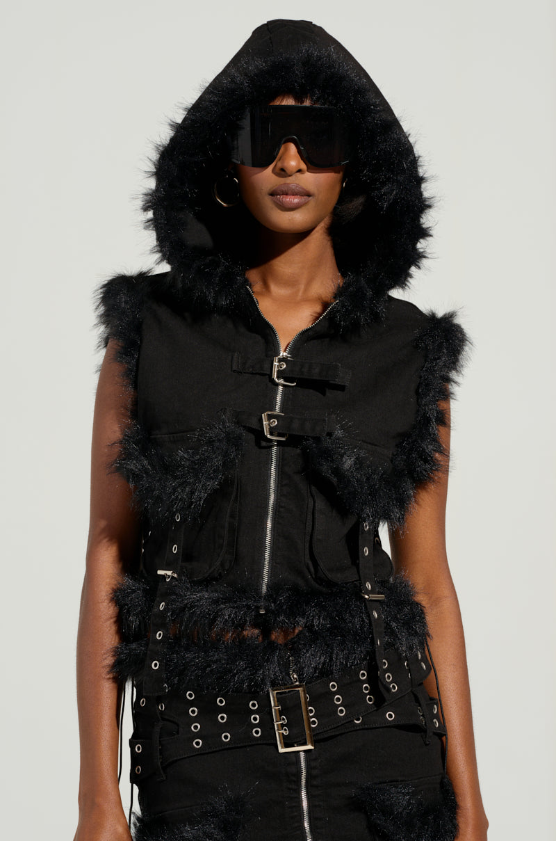 A person flaunting a modern, edgy style in the DELANEY FUR TRIM DENIM VEST, paired with large black sunglasses and a studded belt. The cropped silhouette of the vest accentuates the striking ensemble against the pale backdrop.
