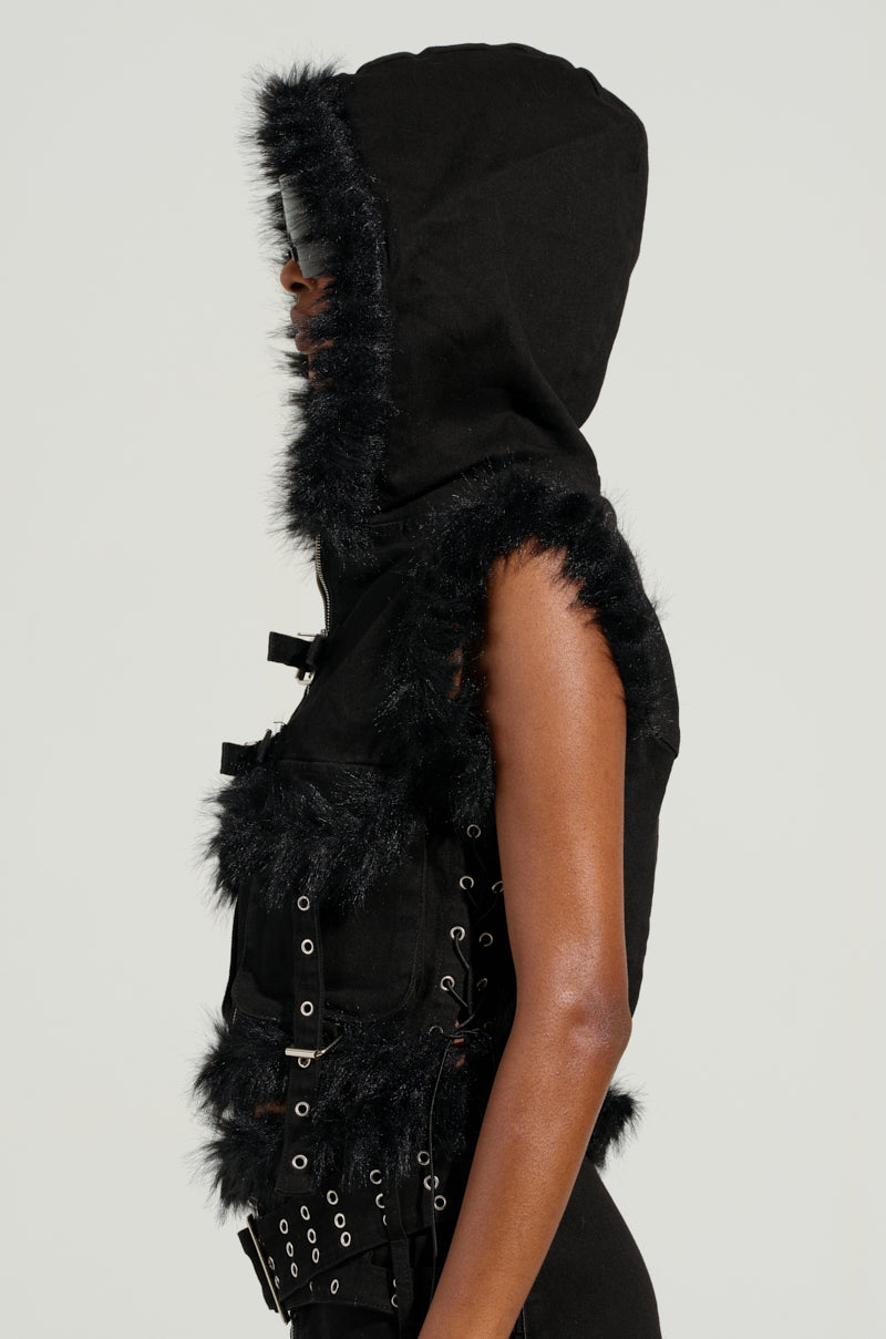 A person wearing the DELANEY FUR TRIM DENIM VEST is displayed from the side, featuring a cropped silhouette with black faux fur trim. The vest is enhanced with decorative straps and buckles.
